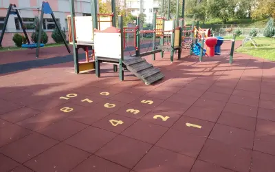 Plac zabaw z huśtawkami i drabinkami. Na czerwonej nawierzchni namalowana jest gra w klasy, a obok niej widać elementy do zabawy w twister.