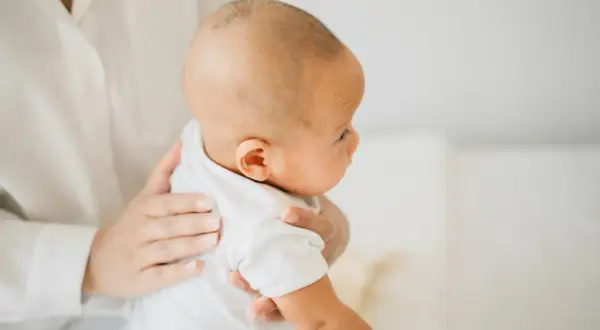 Delikatne dłonie trzymają noworodka w białej koszulce. Mama szuka sposobów, co na czkawkę u noworodka, by ukoić maluszka.