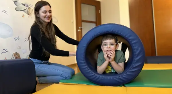 Chłopiec w okularach w tunelu terapeutycznym, obok uśmiechnięta terapeutka.