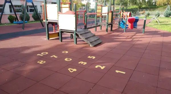 Plac zabaw z huśtawkami i drabinkami. Na czerwonej nawierzchni namalowana jest gra w klasy, a obok niej widać elementy do zabawy w twister.