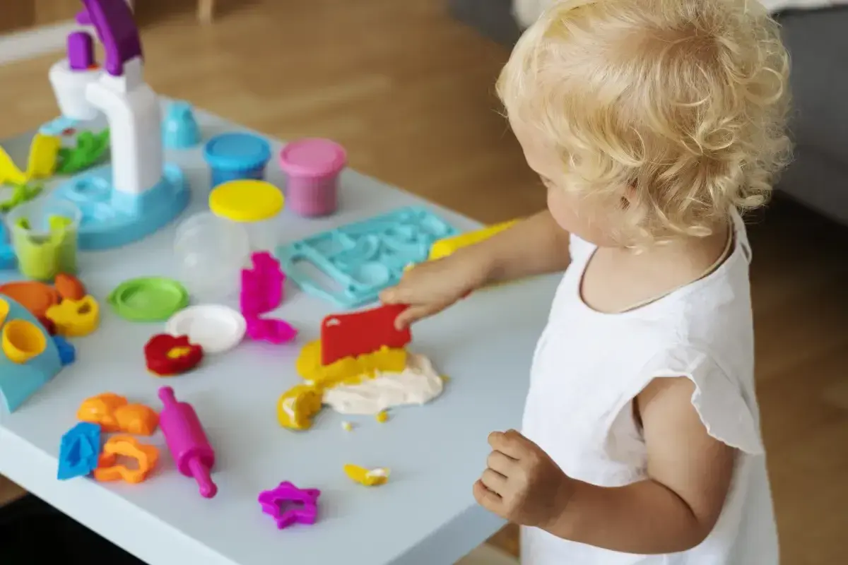 Mała blondynka bawi się masą plastyczną, rozwijając sensorykę. Kolorowe foremki i wałek czekają na kreatywne pomysły.